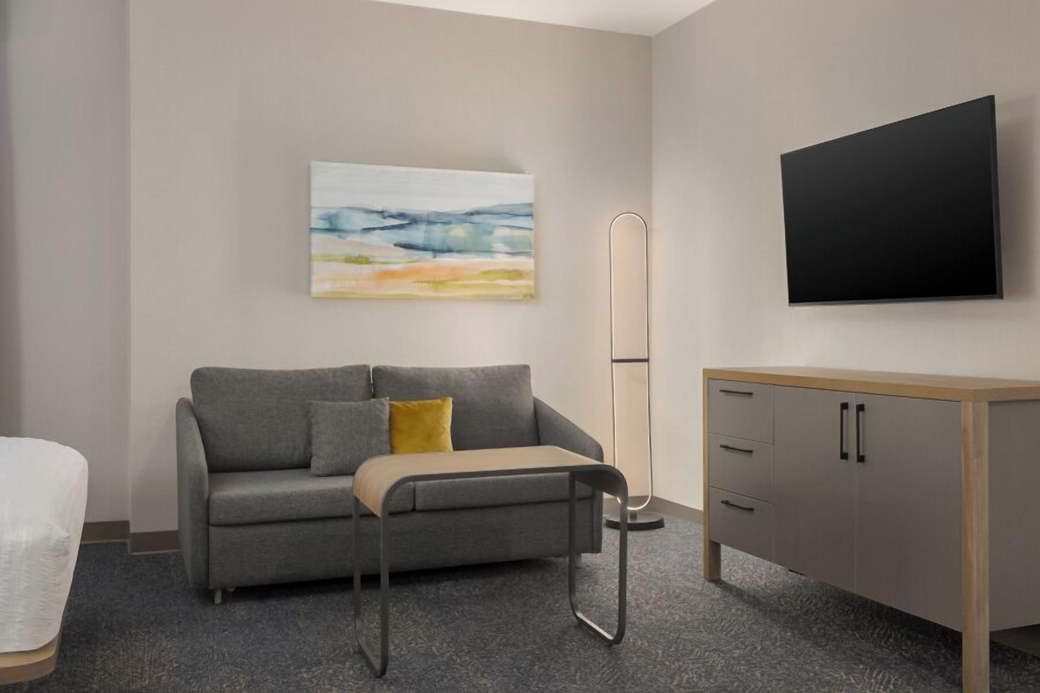 Ein modernes Hotelzimmer mit grauem Sofa, einem kleinen Tisch, einem Holzschrank, einer Stehlampe und einem flachbildfernseher an der Wand.