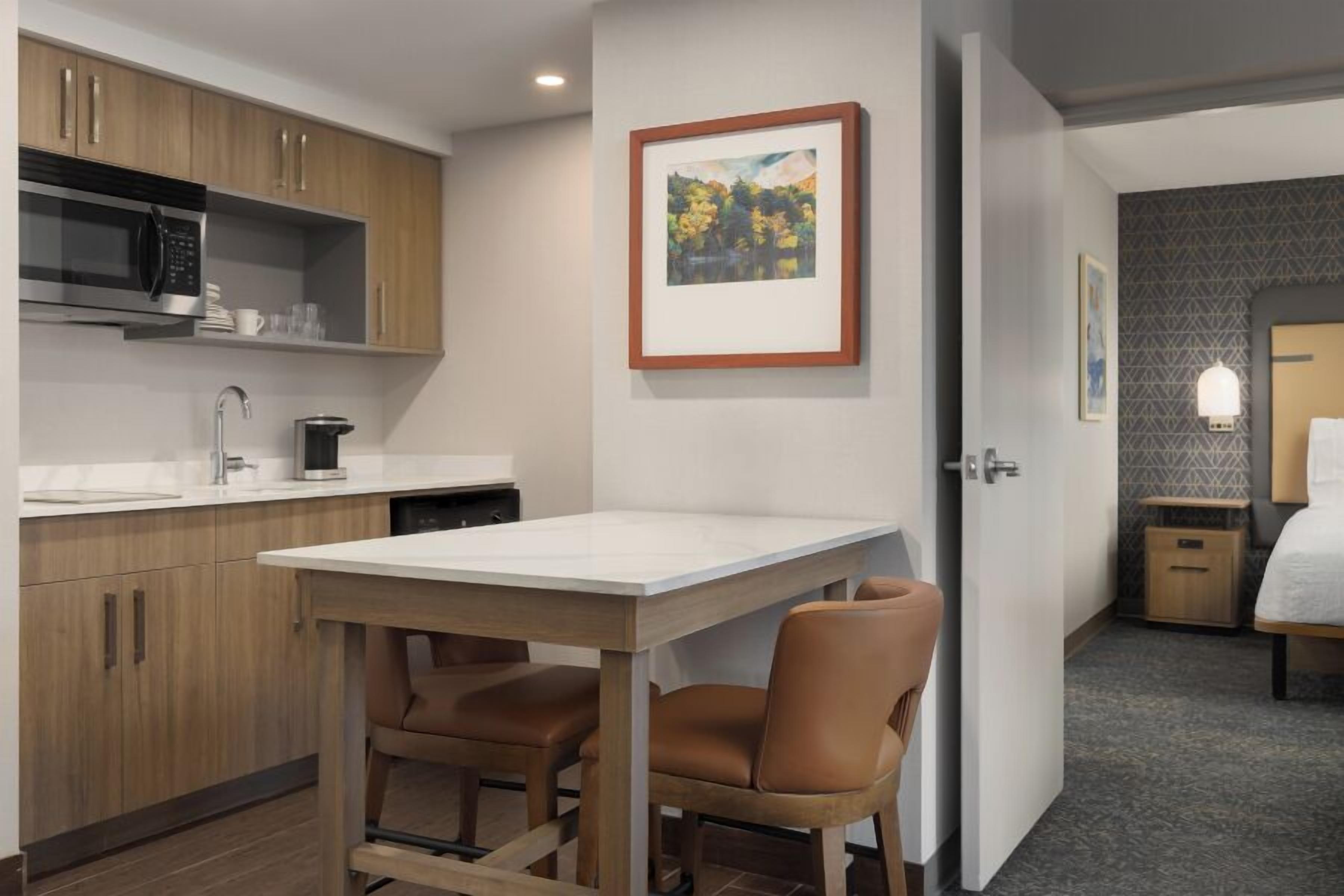Habitación de hotel compacta con kitchenette, zona de comedor con mesa y sillas, y cama con mesita de noche.