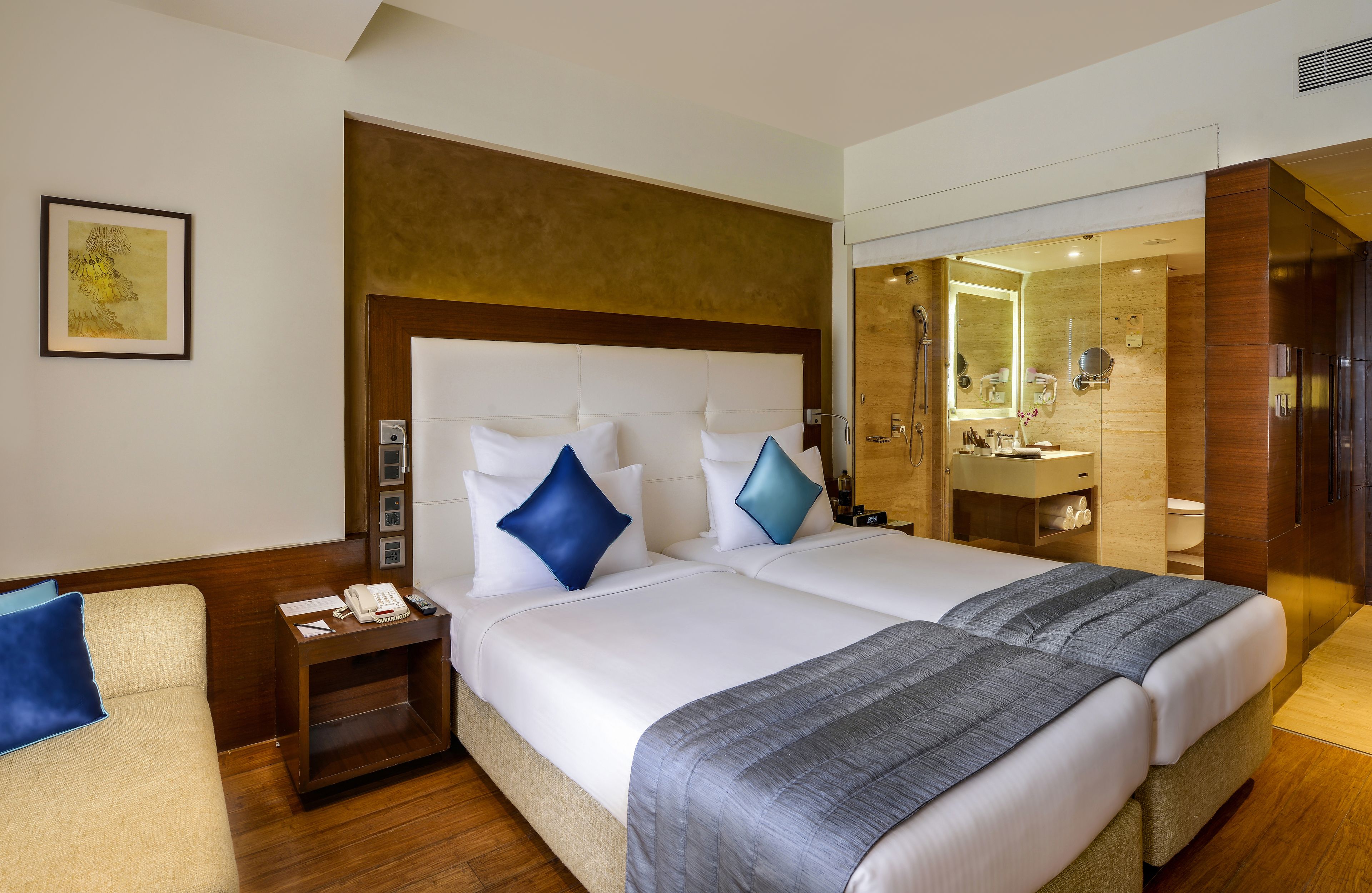 Habitación de hotel con una cama grande, una mesita de noche con teléfono y lámpara, cabecero de madera y un baño con espejo y lavamanos.