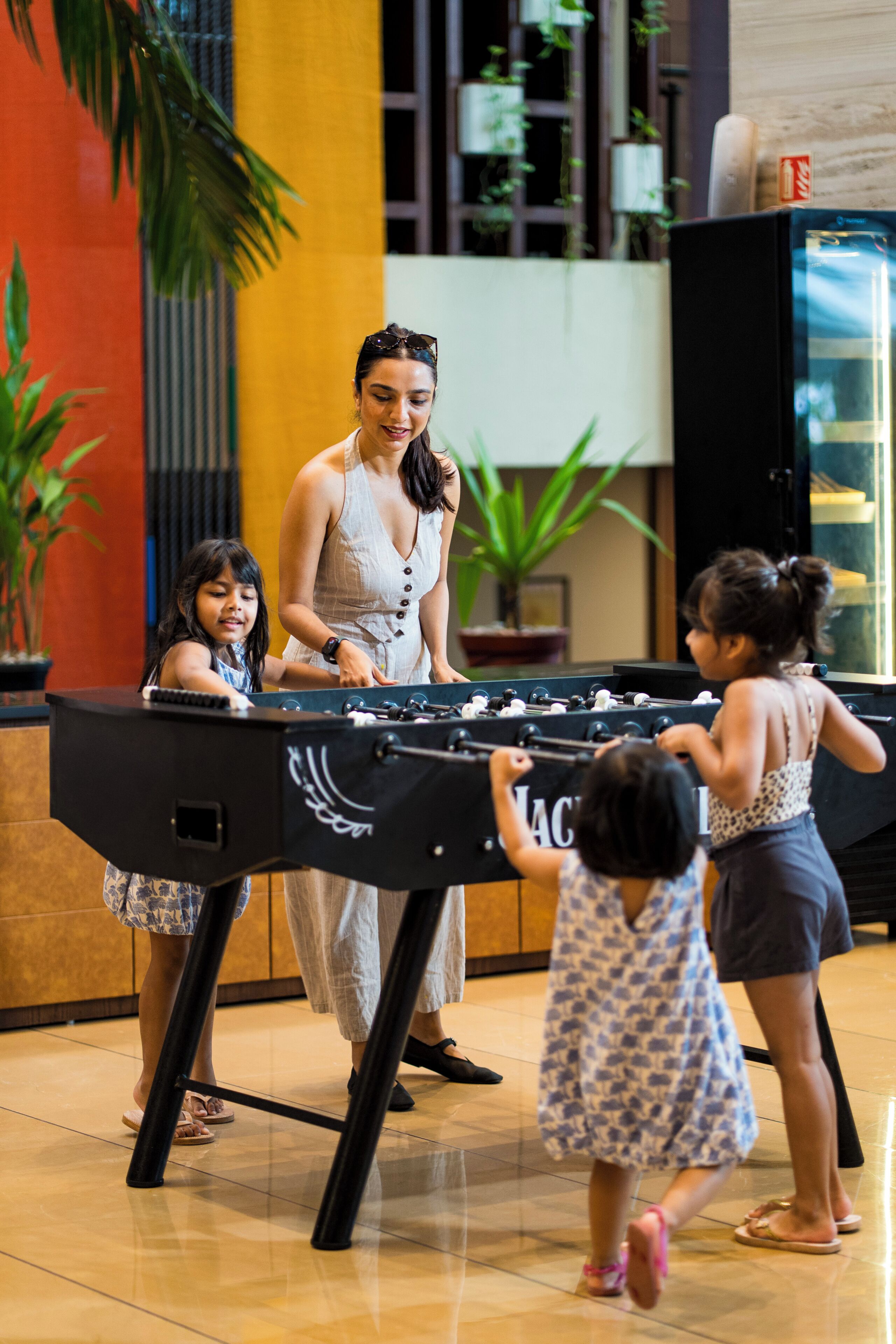 A group of people playing foosball in a room with a large foosball table, potted plants, and a fire extinguisher on the wall.