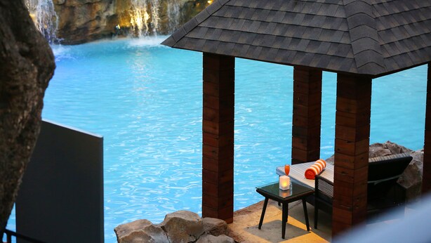 Ein hölzernes Gartenhaus mit einem Ziegeldach, das einen Pool mit einem Wasserfall im Hintergrund überblickt.