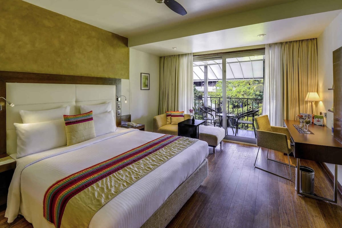 A hotel room with a large bed, a desk, a chair, and a balcony with a view of greenery.