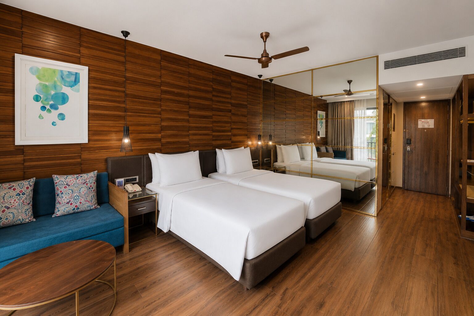 A hotel room with a wooden wall, two beds, a sofa, a desk, and a ceiling fan.