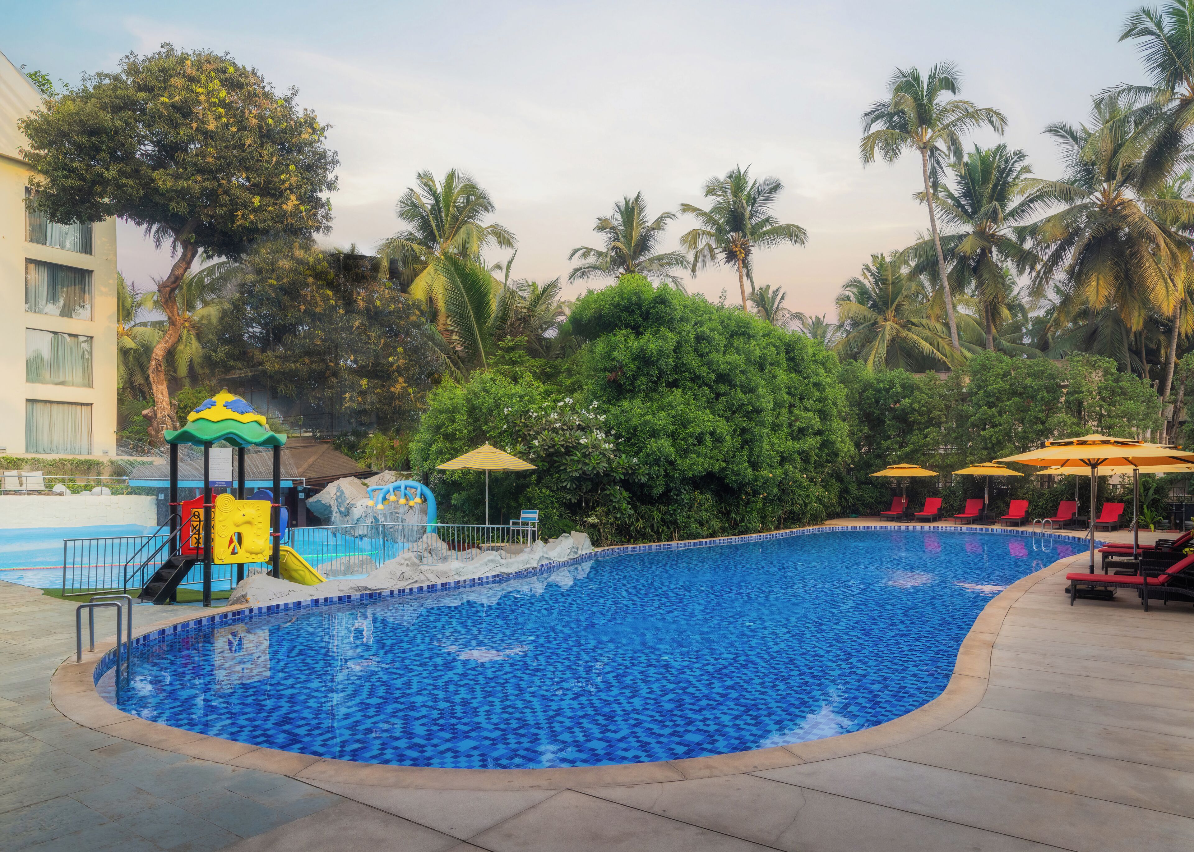 Une piscine avec un espace de jeux pour enfants, des palmiers et des chaises longues.