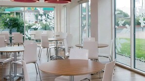 A spacious indoor dining area with round tables and white chairs, large windows offering a view of the outdoors, and red circular ceiling lights. - Travelodge Manchester Piccadilly (Manchester)