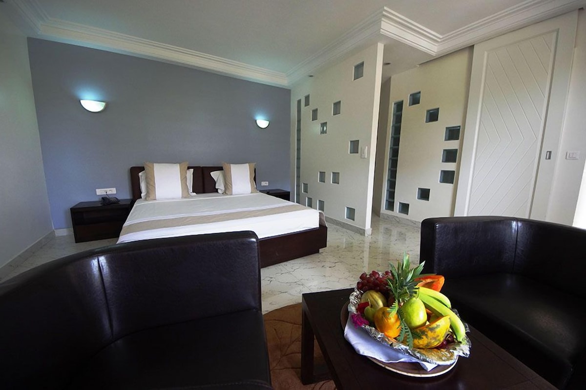 Een hotelkamer met een bed, twee nachtkastjes, een salontafel met fruit en een zwarte leren bank.