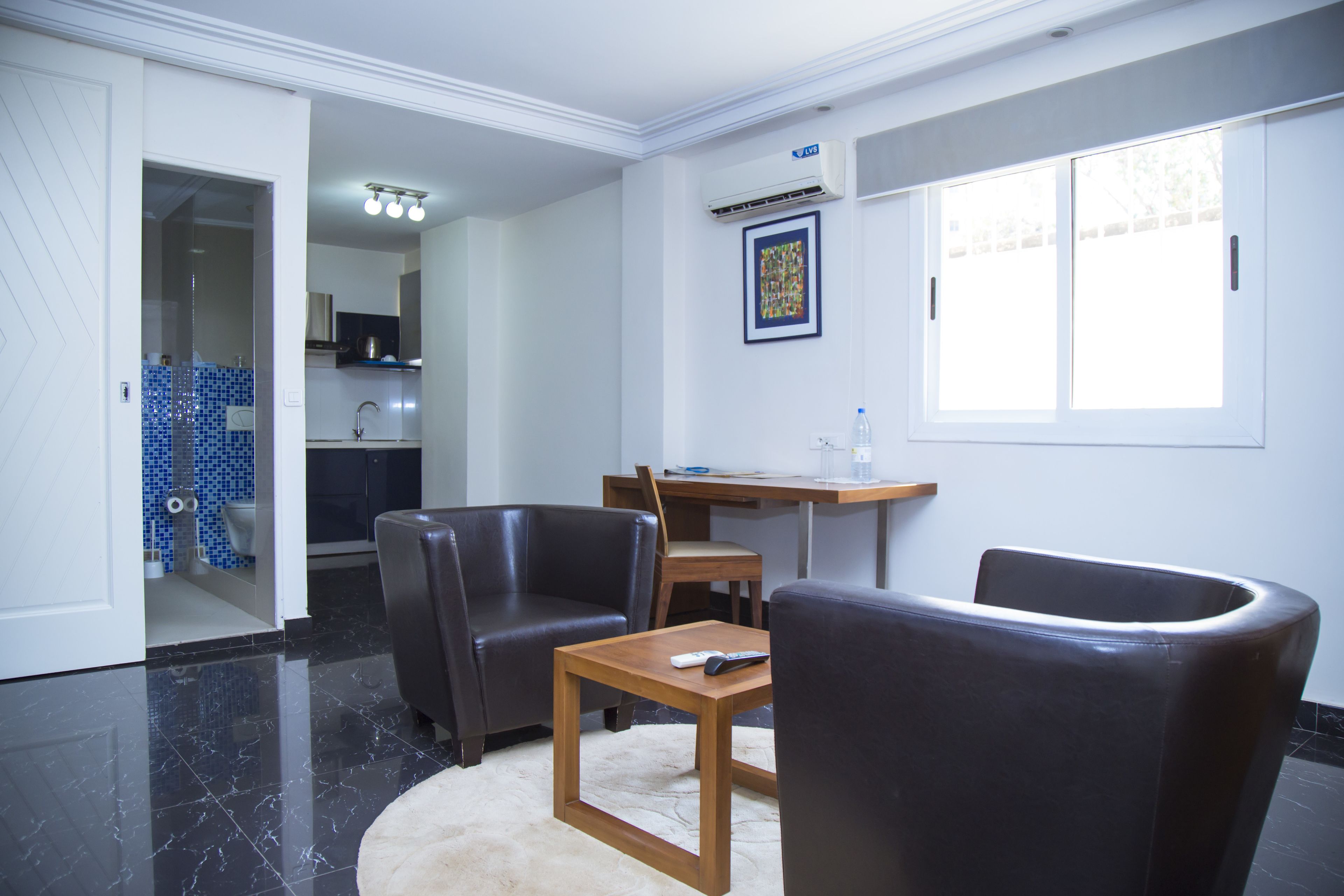 A modern living room with a black leather sofa, a wooden coffee table, and a remote control on it. There is a dining table with chairs, a kitchen area, and a bathroom visible through an open door.
