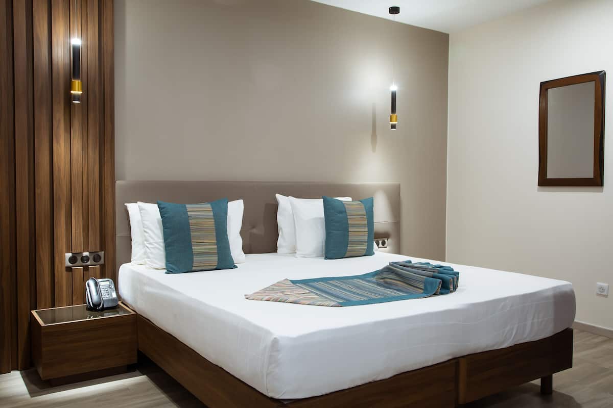 Una cama bien hecha con sábanas blancas y almohadas color turquesa, una mesita de noche de madera con un teléfono, y un aplique en la pared.