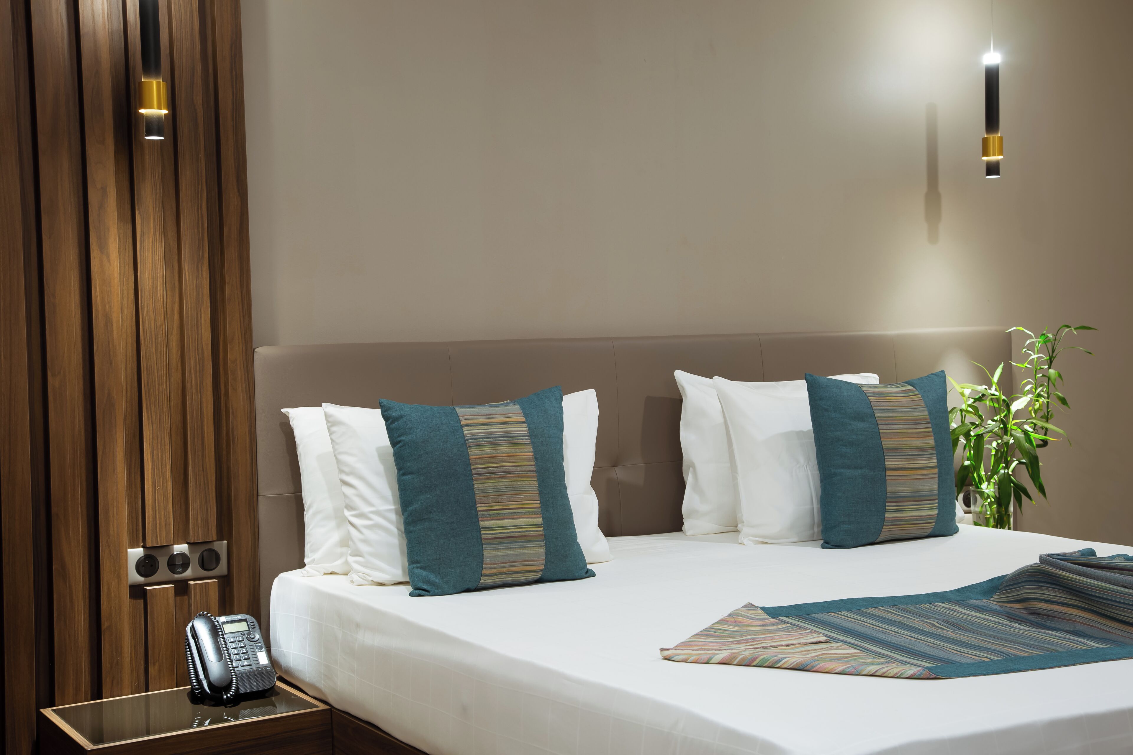 A hotel room with a bed, pillows, a bedside table with a phone, a wall-mounted light, and a potted plant.