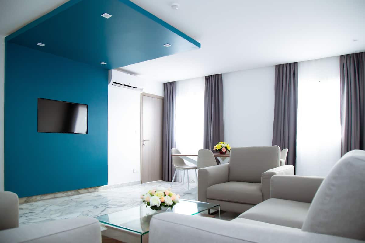 Una sala de estar moderna con una pared azul de acento, un televisor de pantalla plana, una mesa de centro de vidrio con flores y una zona de estar con un sofá y sillones.