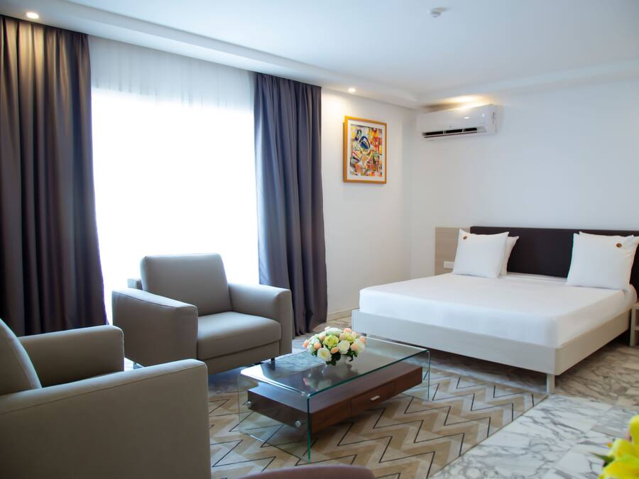 Habitación de hotel con cama, dos sillones, una mesa de centro de cristal con flores, un cuadro en la pared y un equipo de aire acondicionado.