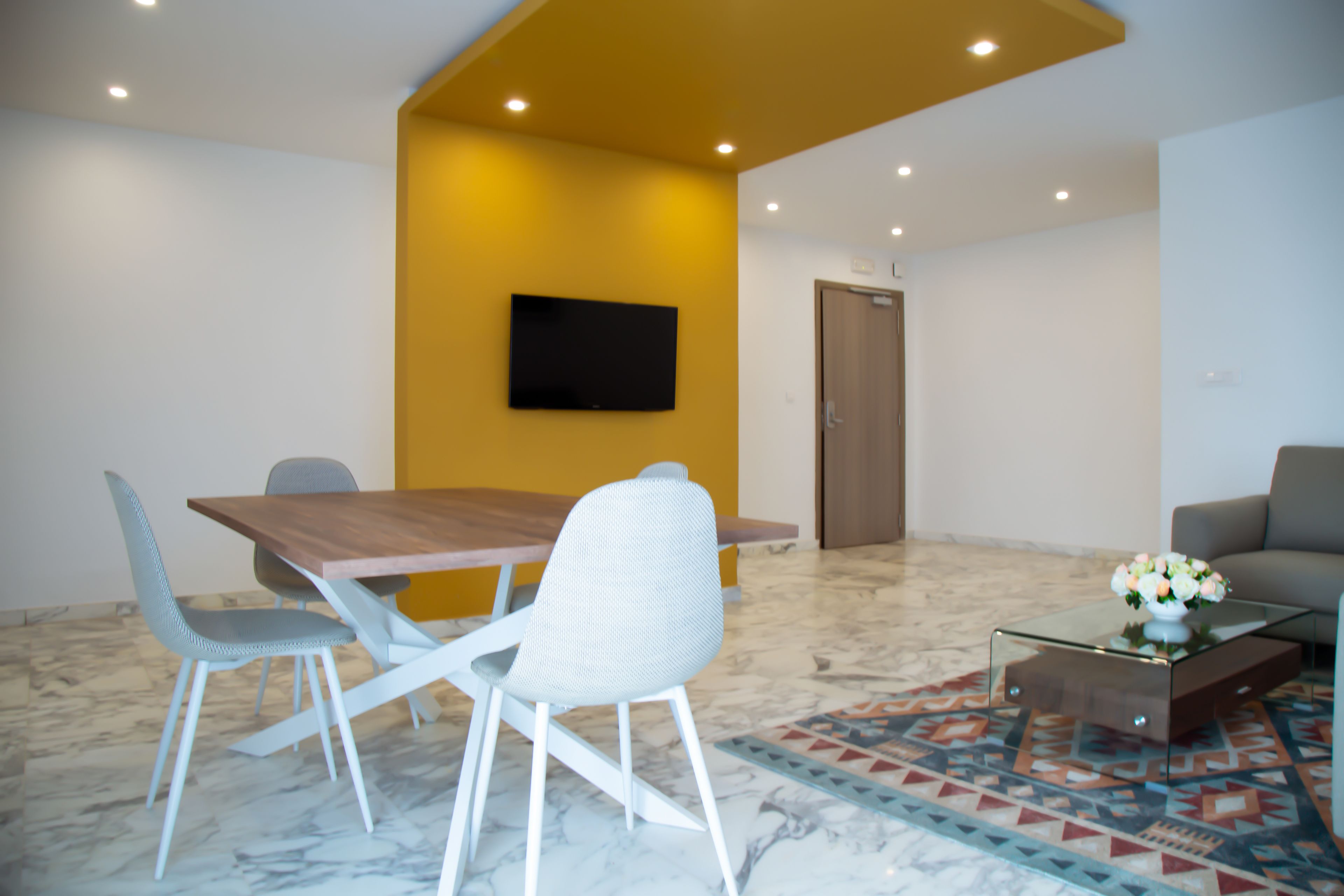 Una sala de reuniones moderna con una mesa de madera, sillas blancas, una mesa baja de vidrio con flores y un televisor de pantalla plana en la pared.