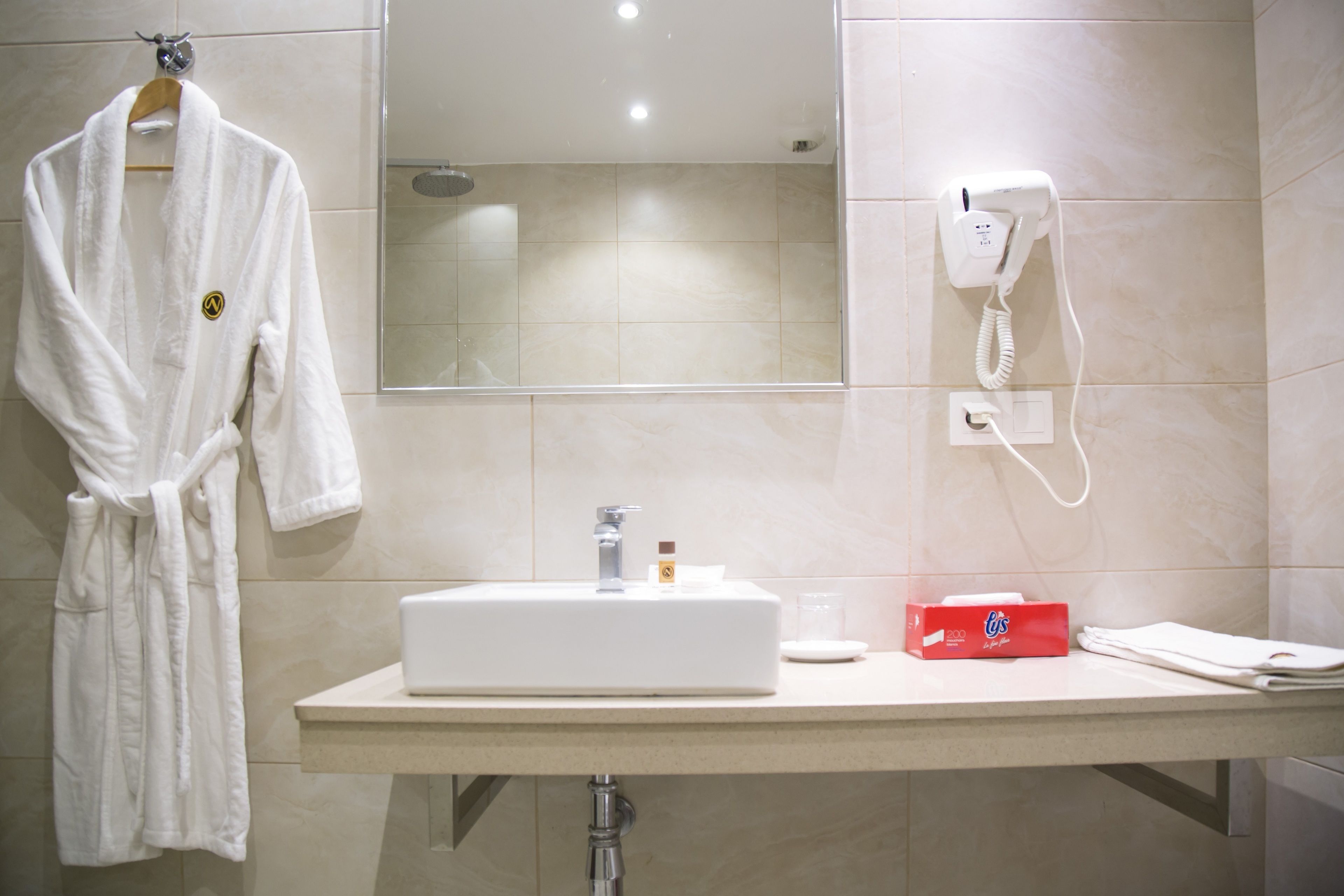 Un baño con un lavabo blanco, un espejo, una bata colgada en un perchero y un secador de pelo en la pared.