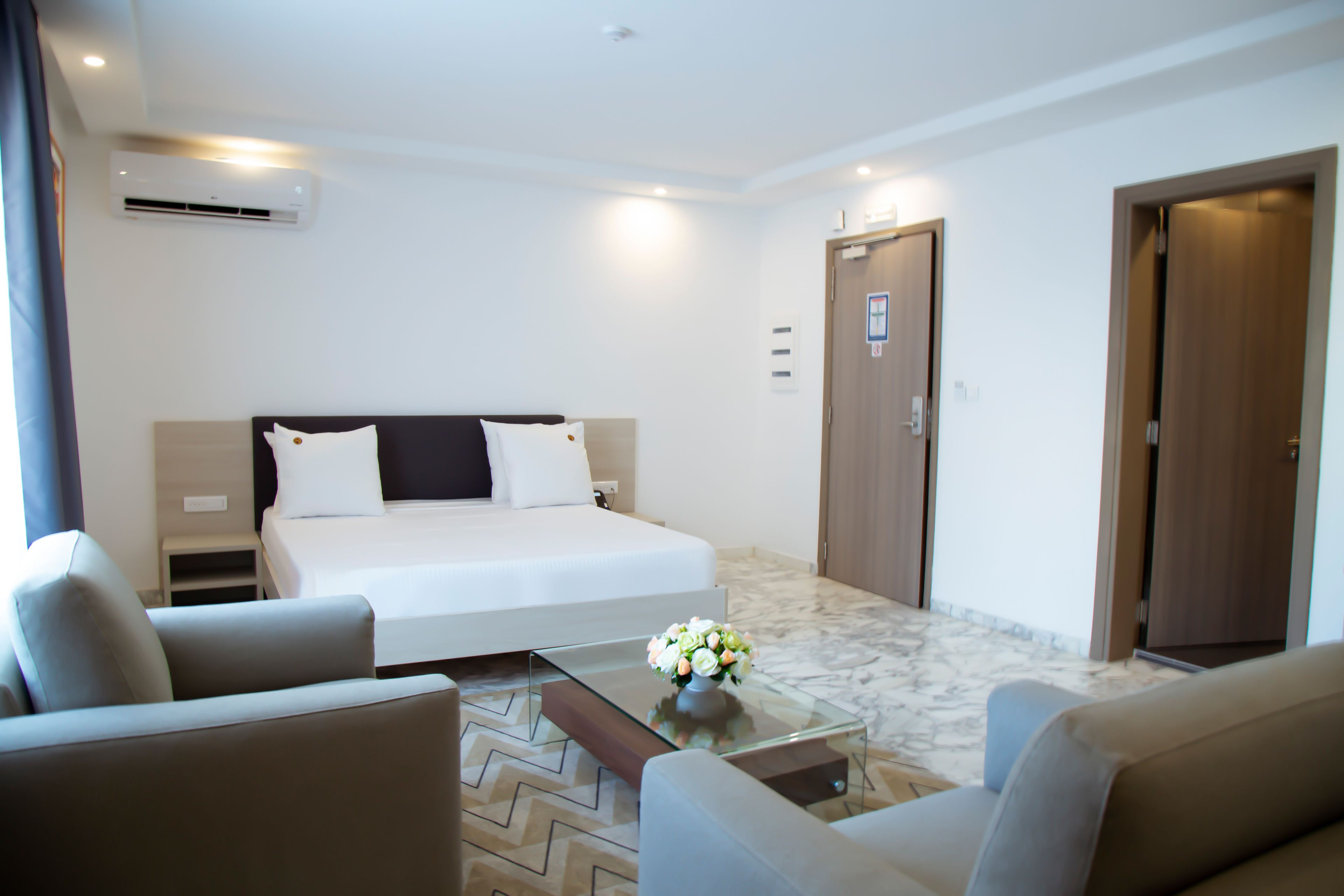 Habitación de hotel con cama, una mesa de centro de vidrio con flores, un sofá y piso alfombrado.