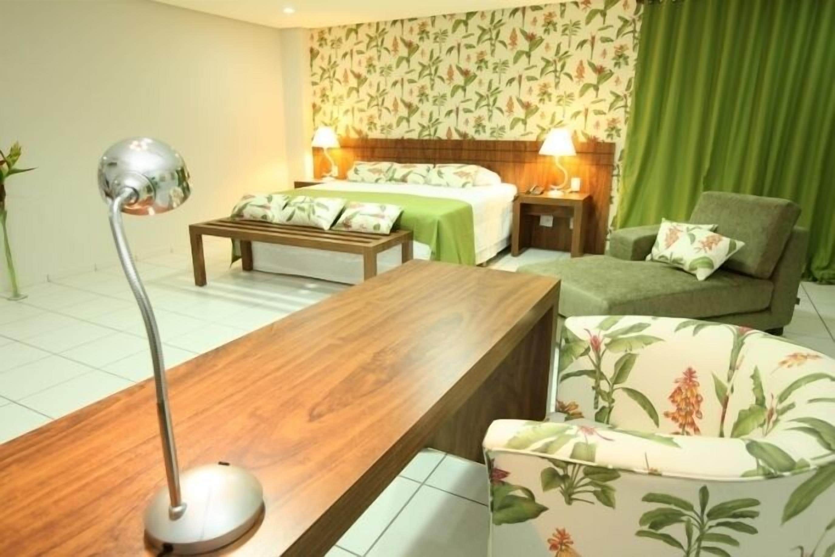A hotel room with a wooden desk, a green armchair, a bed with a green bedspread, and a floral wallpaper.