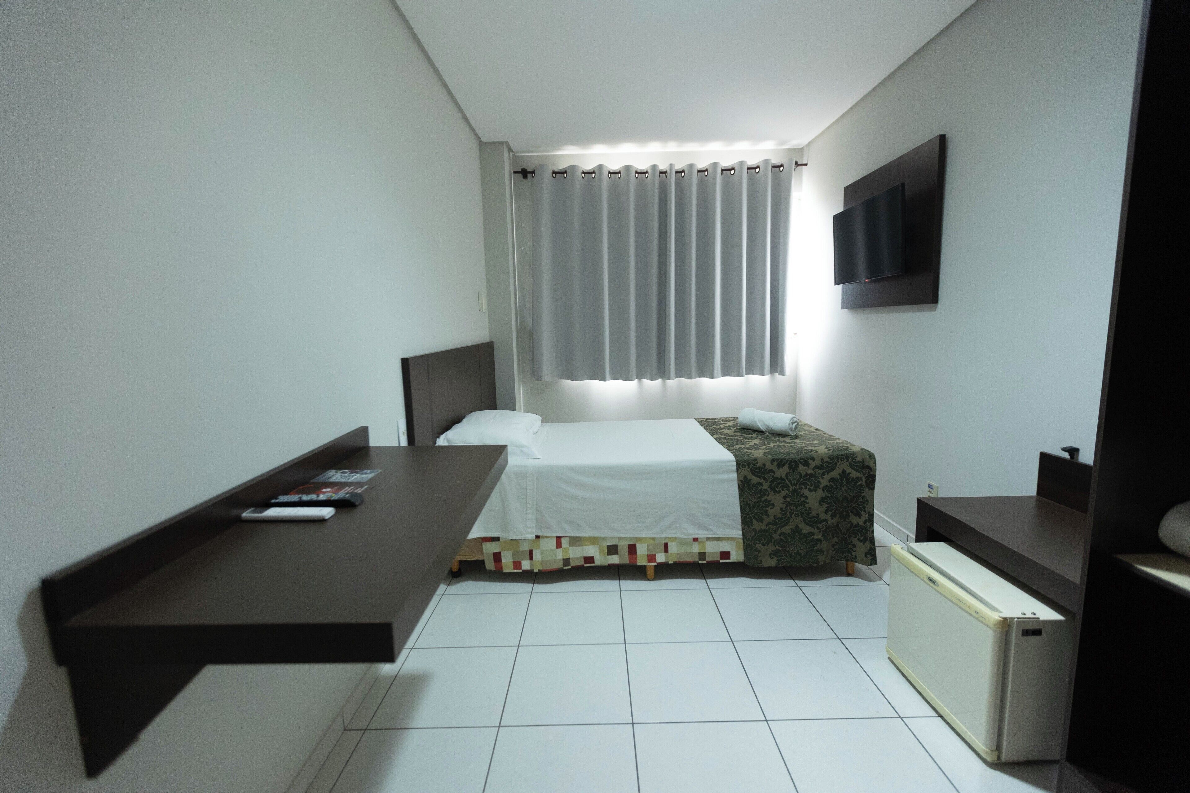 Una camera da letto piccola e ordinata, con un letto, un comodino, una TV fissata al muro e una finestra con tende.