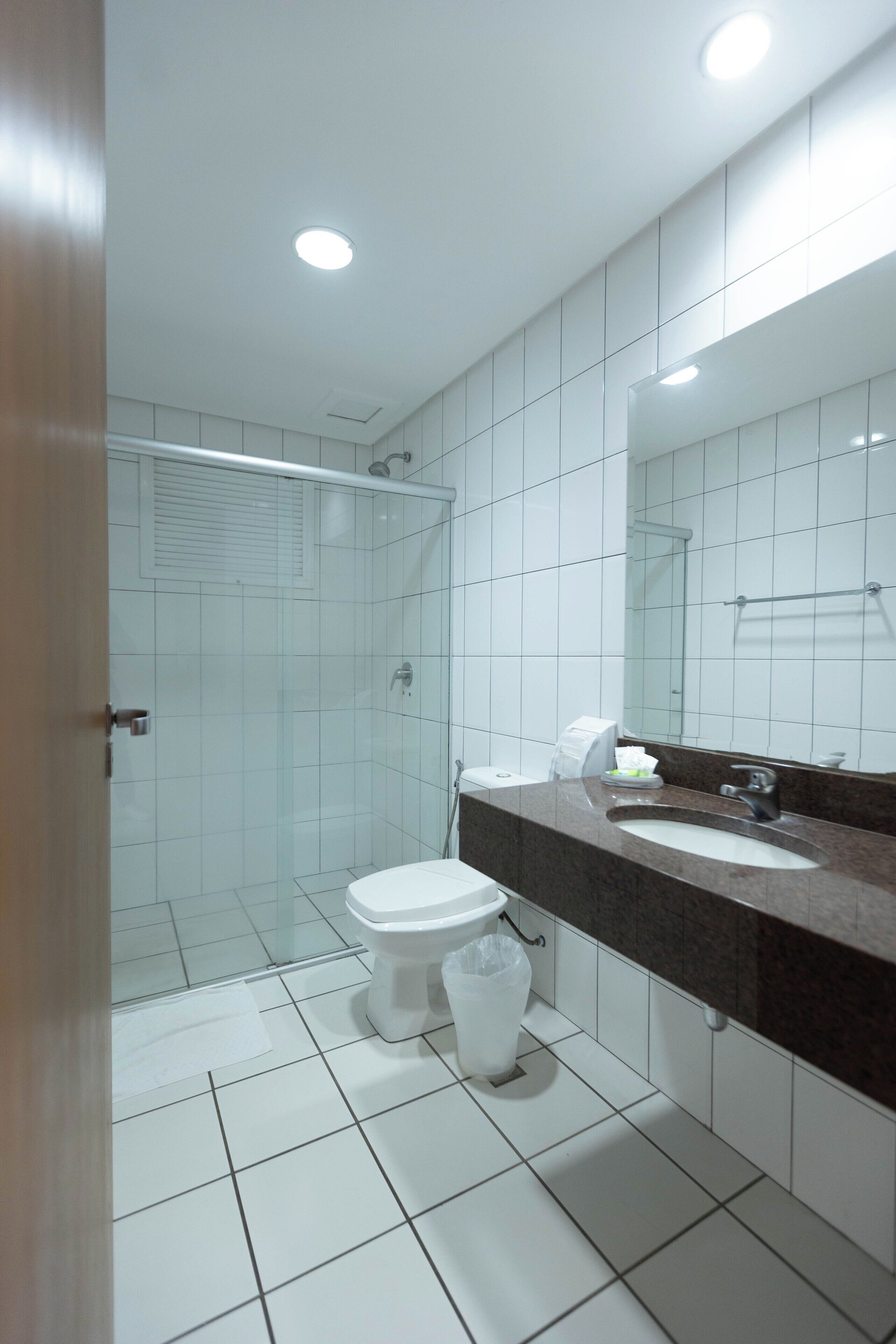 A modern bathroom with white tiles, a glass shower enclosure, a white toilet, and a dark brown countertop with a sink and mirror.