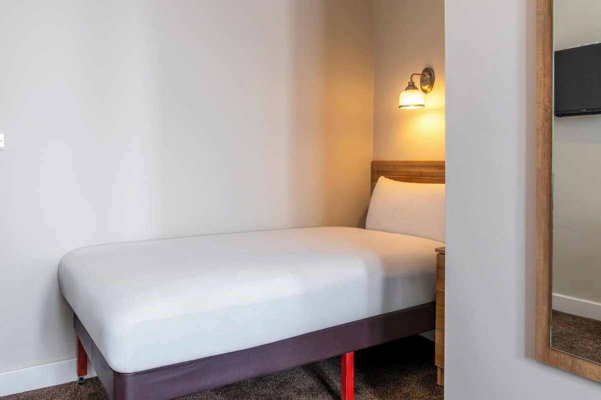 Un lit simple avec une tête de lit en bois, une table de chevet et un luminaire fixé au mur.