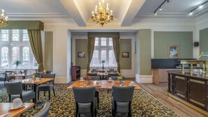Een restaurantgedeelte van een hotel, met een grote tafel, stoelen, een kroonluchter en een vloerkleed.