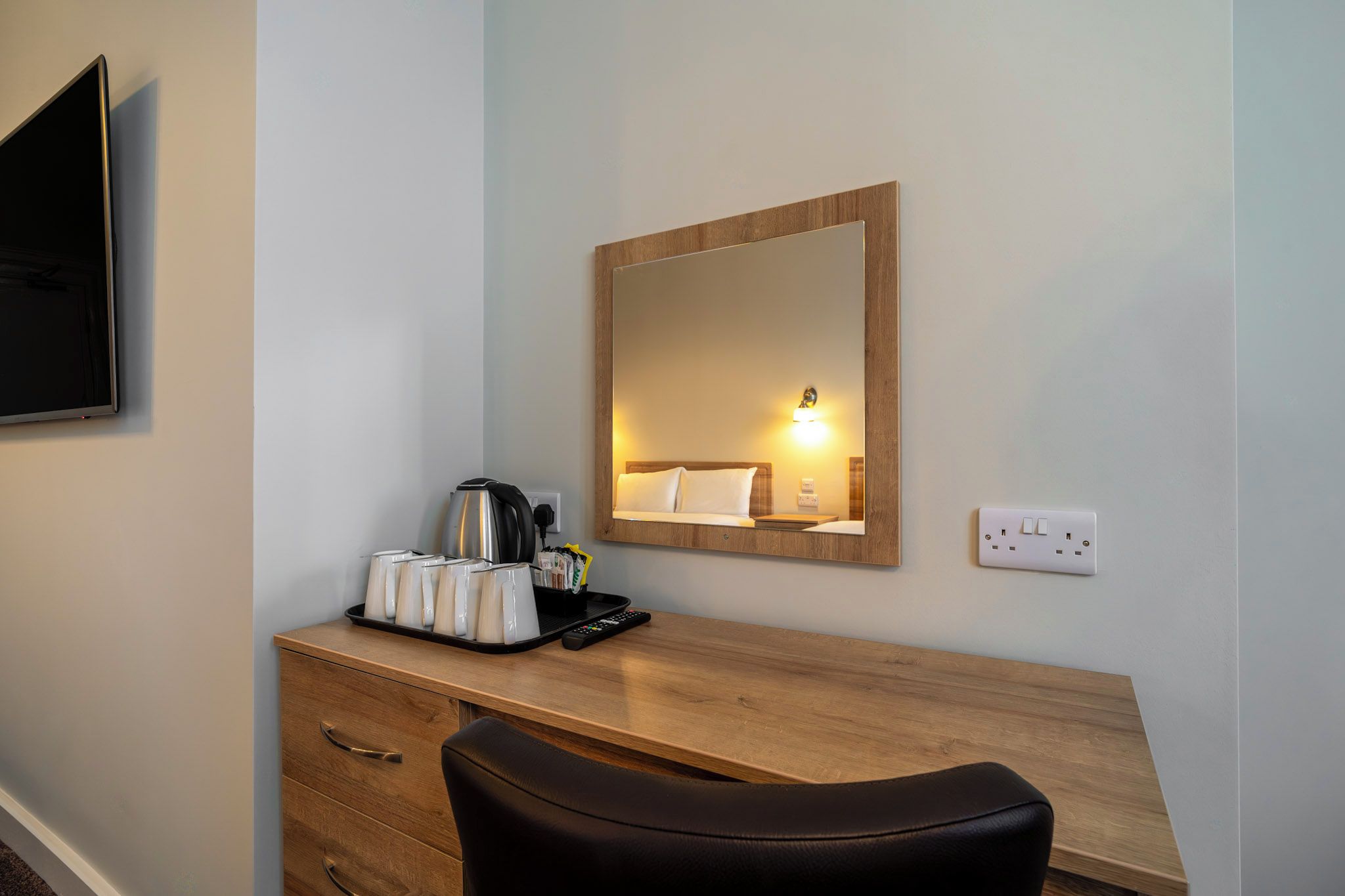 A hotel room with a wooden desk, a black leather chair, a coffee maker, a tray with cups and a remote control, and a mirror on the wall.