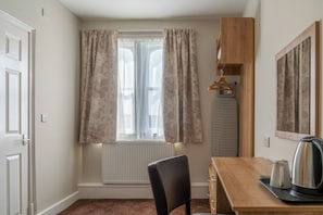Une pièce comprenant un bureau en bois, une chaise, un radiateur, une fen être avec des rideaux, un porte-manteau et une bouilloire.