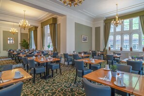 Une salle à manger d’hôtel avec de nombreuses tables, des chaises, des lustres et de grandes fenêtres.