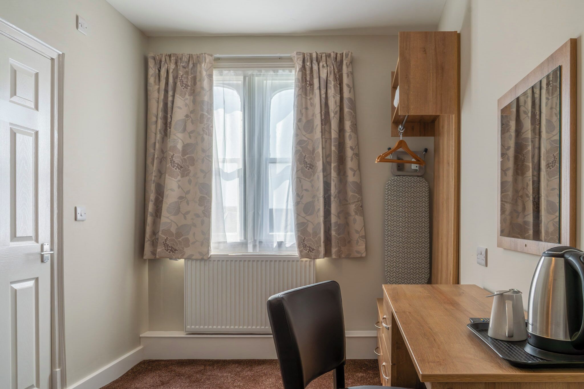Una habitación con un escritorio de madera, una silla, un radiador, una ventana con cortinas, un perchero y una tetera.