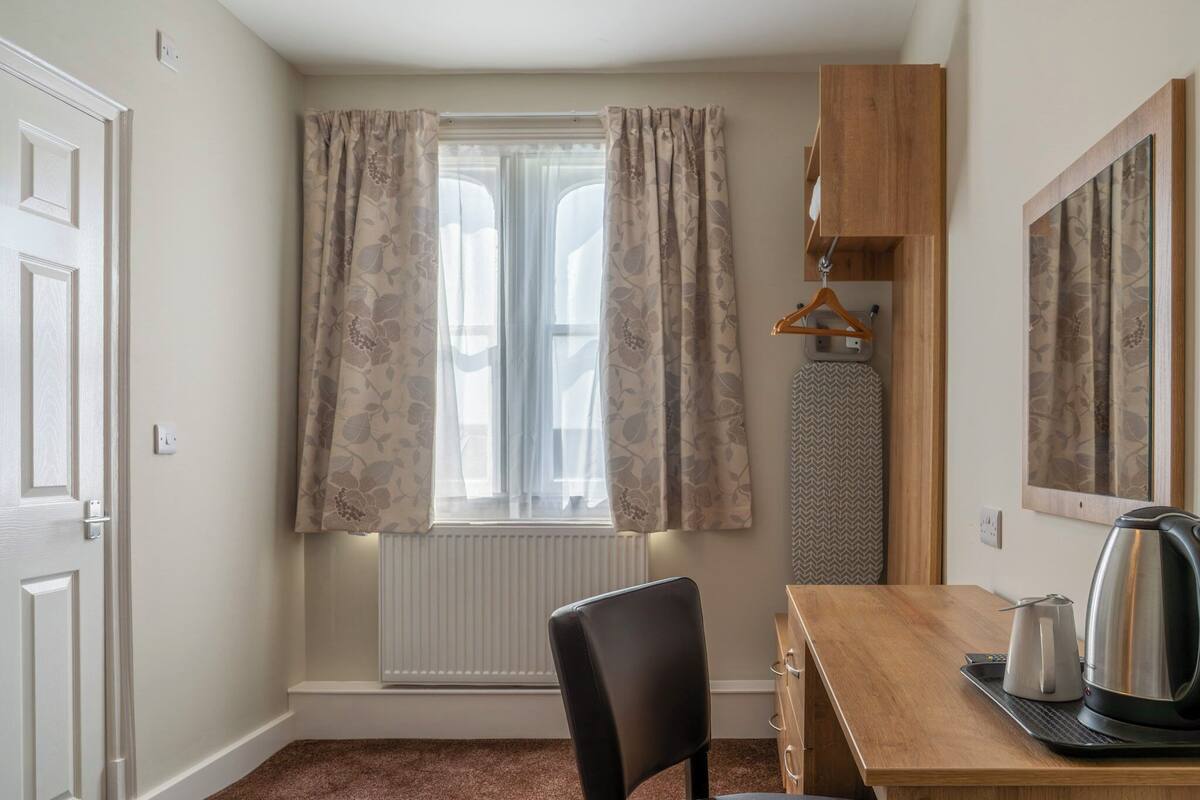 Ein Zimmer mit Holztisch, Stuhl, Heizkörper, einem fenster mit Vorhängen, einer Kleiderstange und einem Wasserkocher.