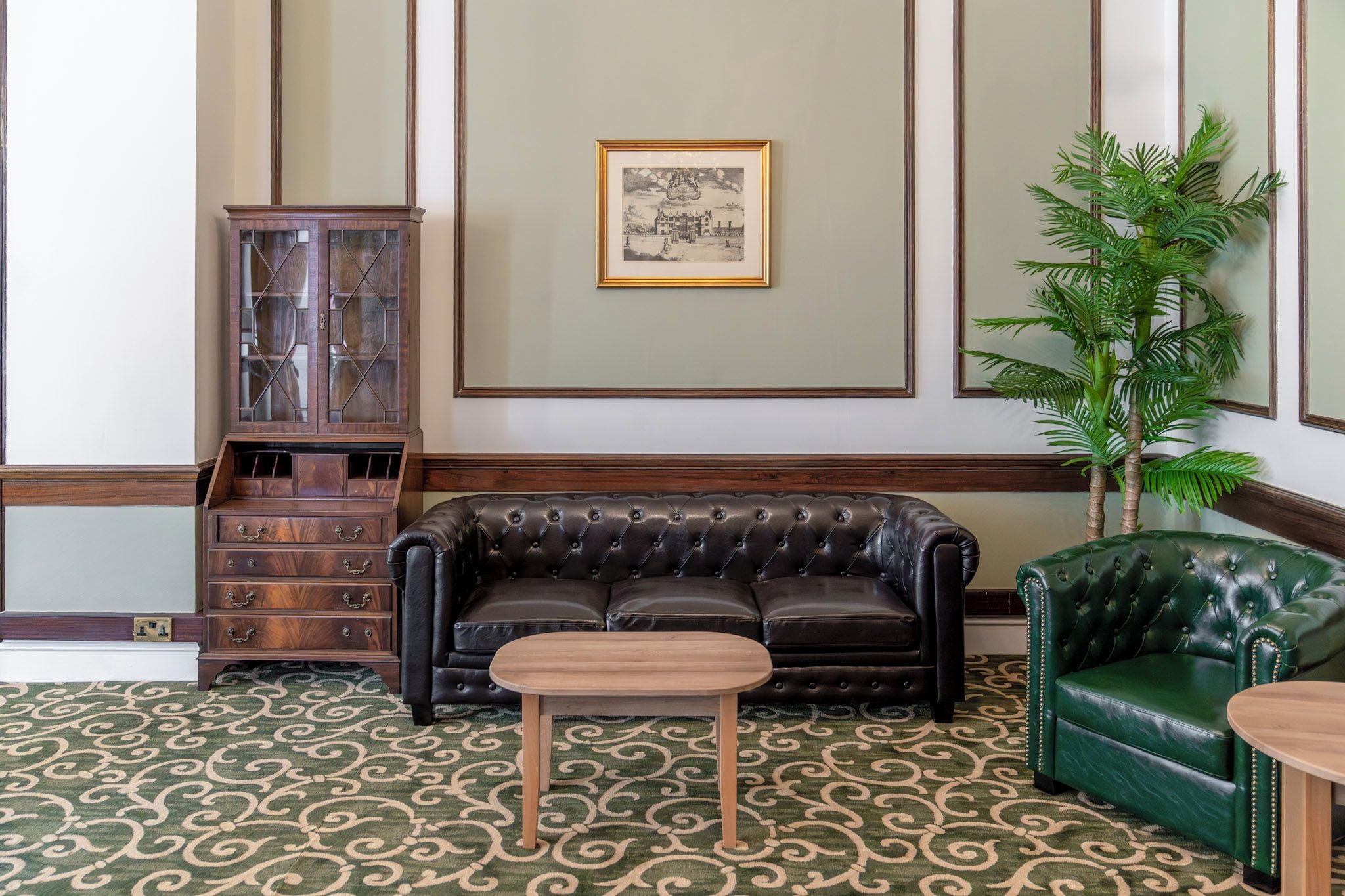A room with a patterned carpet, a black leather sofa, a green armchair, a wooden cabinet, and a framed picture on the wall.