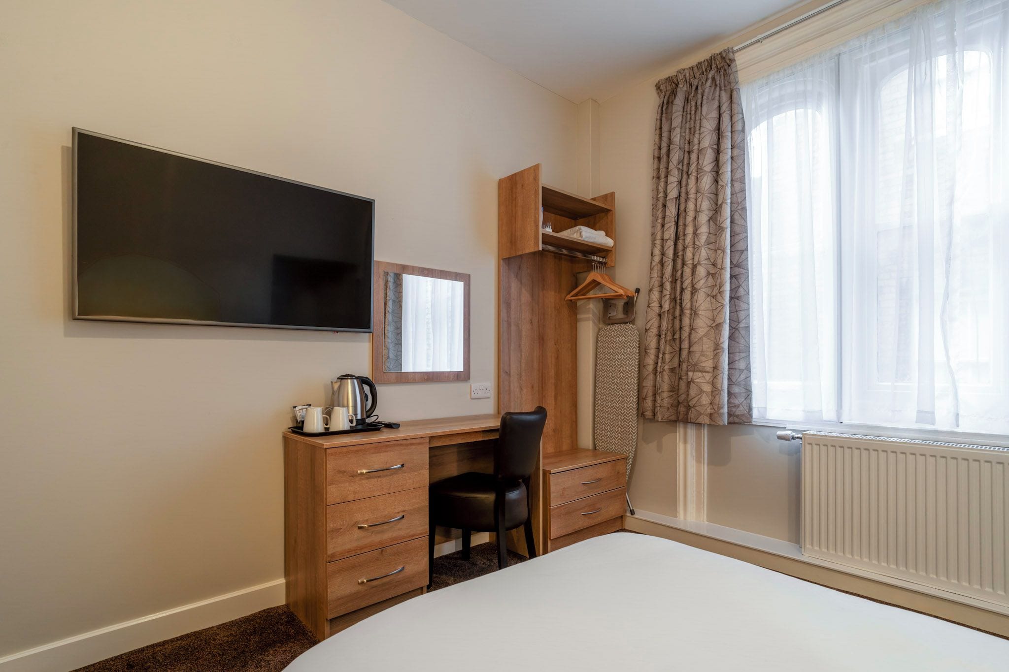 A bedroom with a bed, a desk with a chair, a TV, a wardrobe, and a window with curtains.