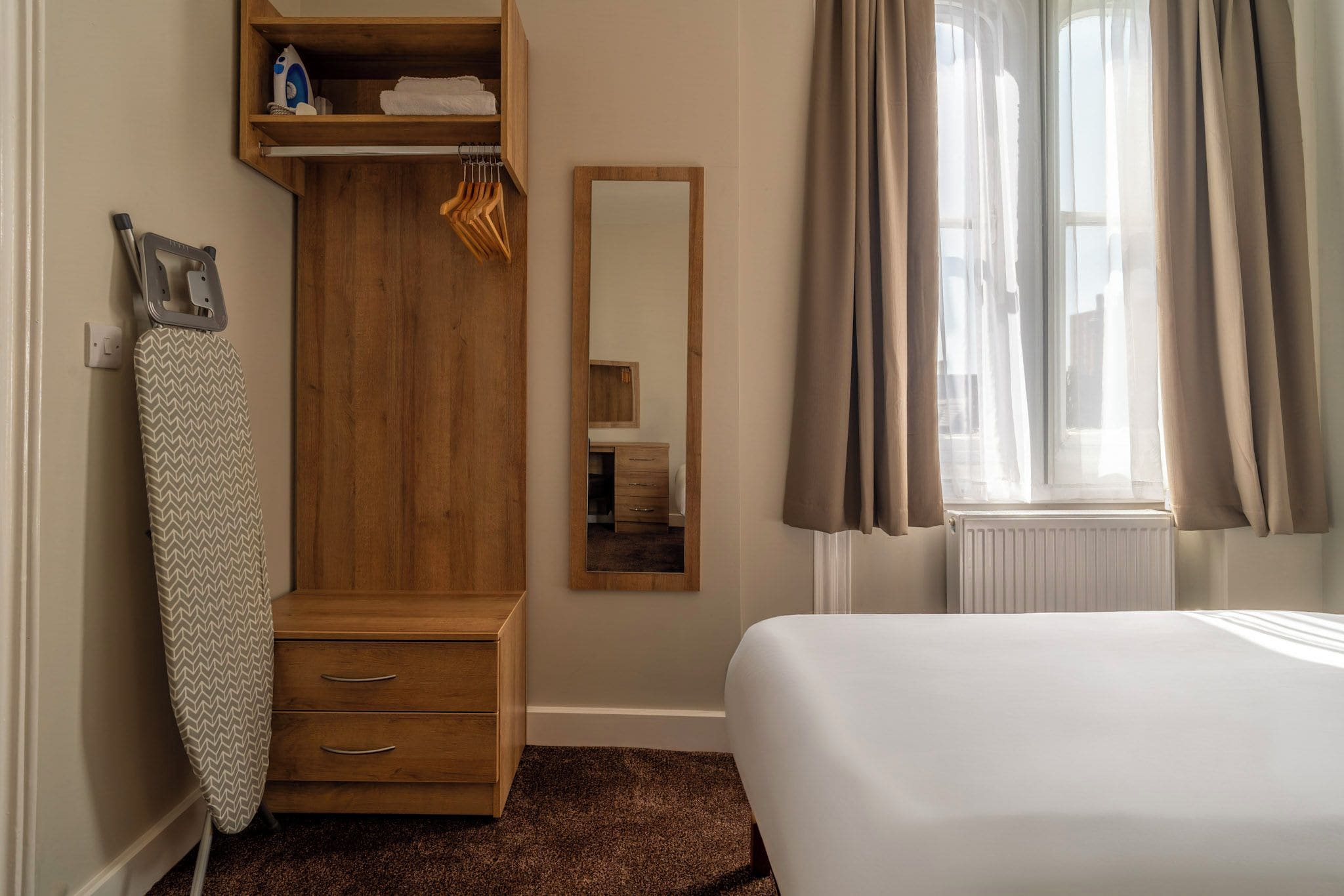 Een slaapkamer met een houten garderobe, een spiegel, een bed en een raam met gordijnen.