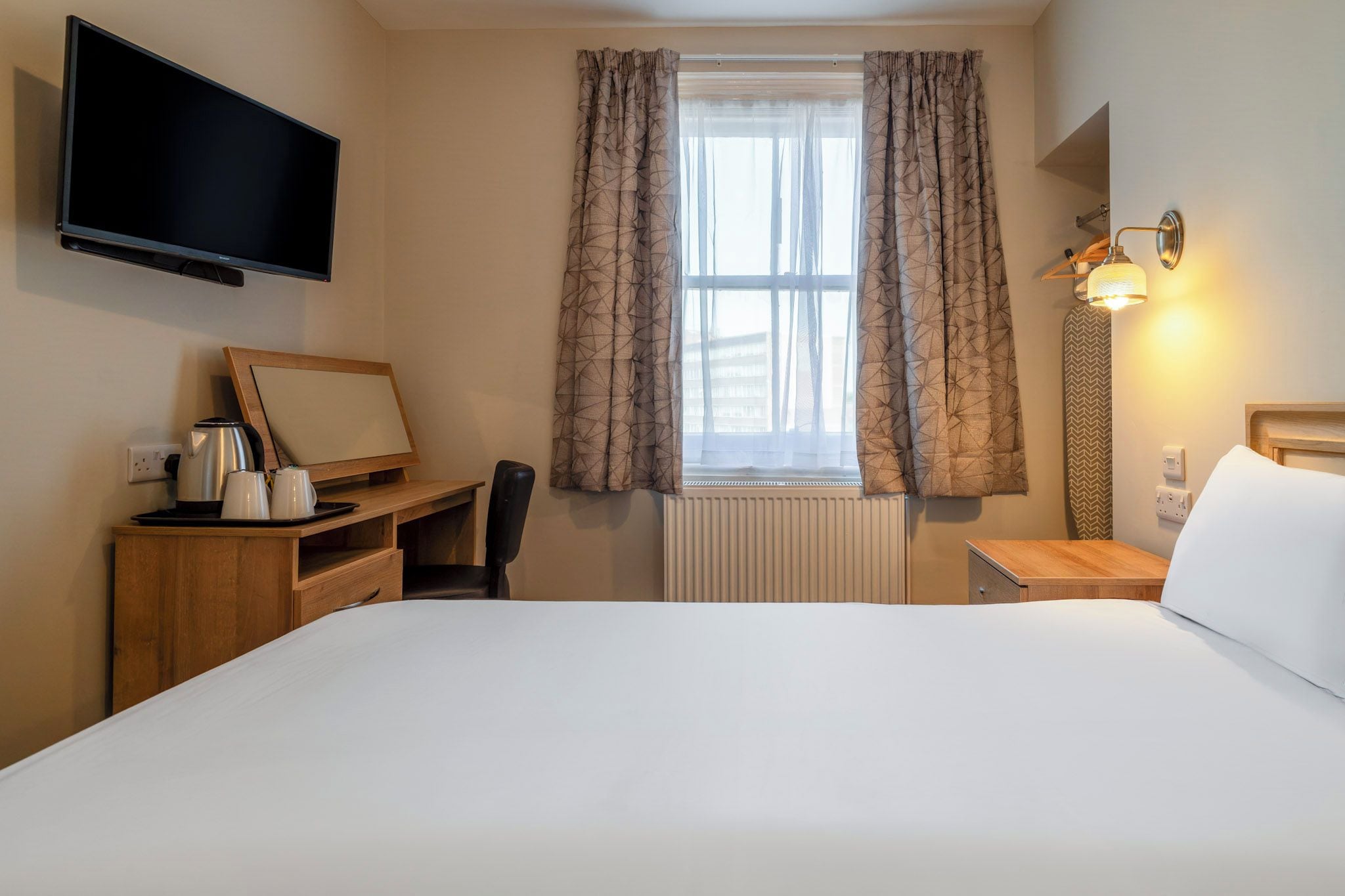Habitación de hotel con cama, escritorio con silla, televisión, ventana con cortinas y lámpara de noche.
