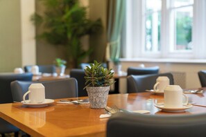 Un coin repas avec des tables préparées pour deux personnes, chacune agrémentée d’une tasse et d’une soucoupe blanches, d’une petite plante en pot et d’une fenêtre avec des rideaux.