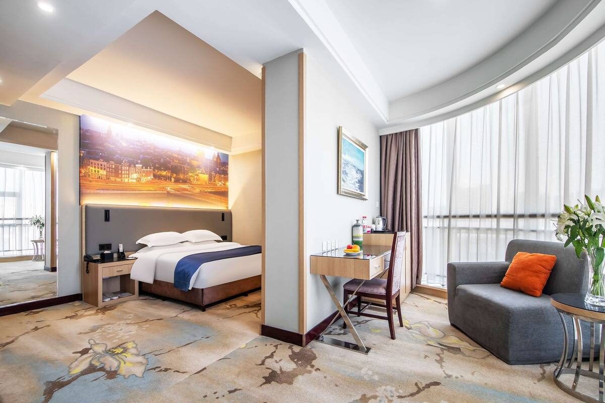 Una habitación de hotel con cama, escritorio, silla y un ventanal con cortinas.