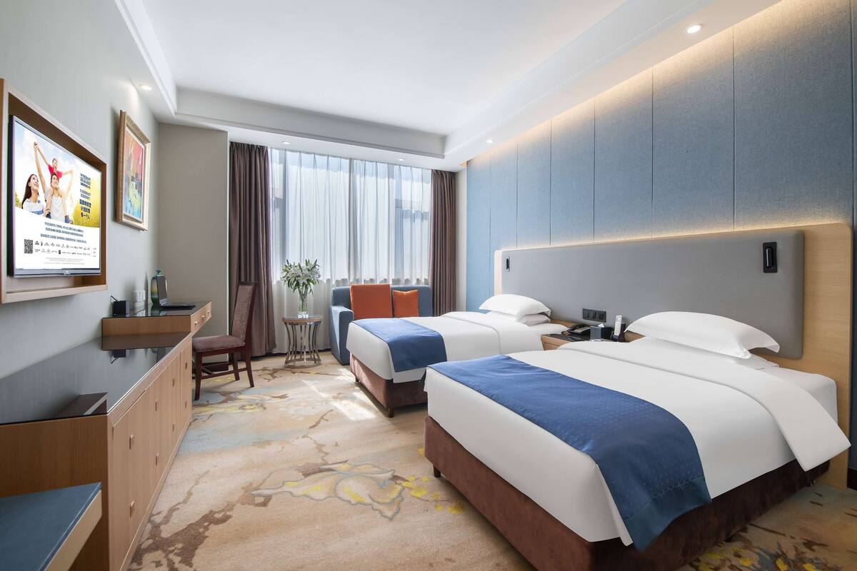 Habitación de hotel con dos camas, un escritorio, una silla y un amplio ventanal con cortinas.