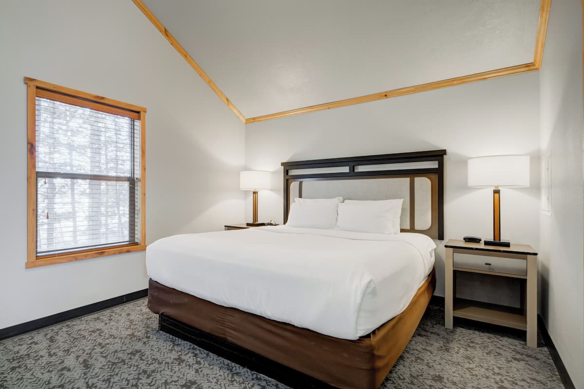 Un cuarto de hotel ordenado con una cama grande, dos mesitas de noche con lámparas, una ventana con persianas y piso alfombrado.