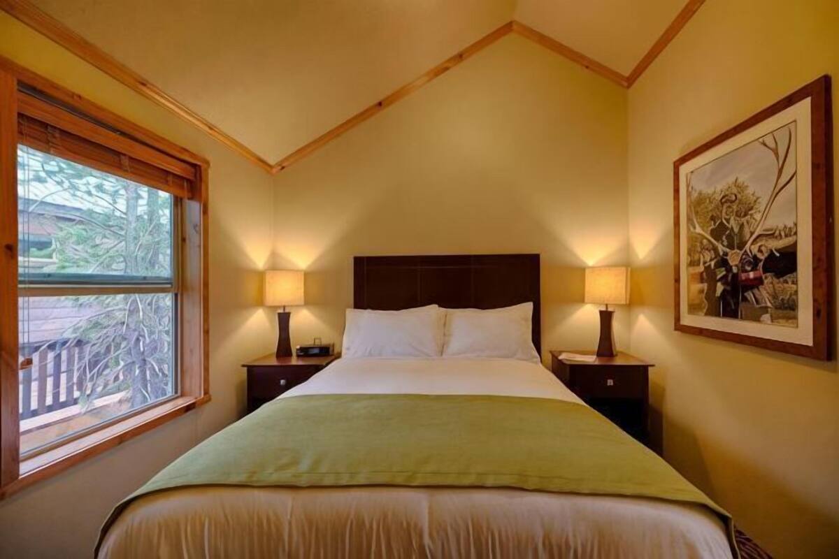 A bedroom with a large bed, two bedside tables with lamps, a window with a view, and a framed picture on the wall.