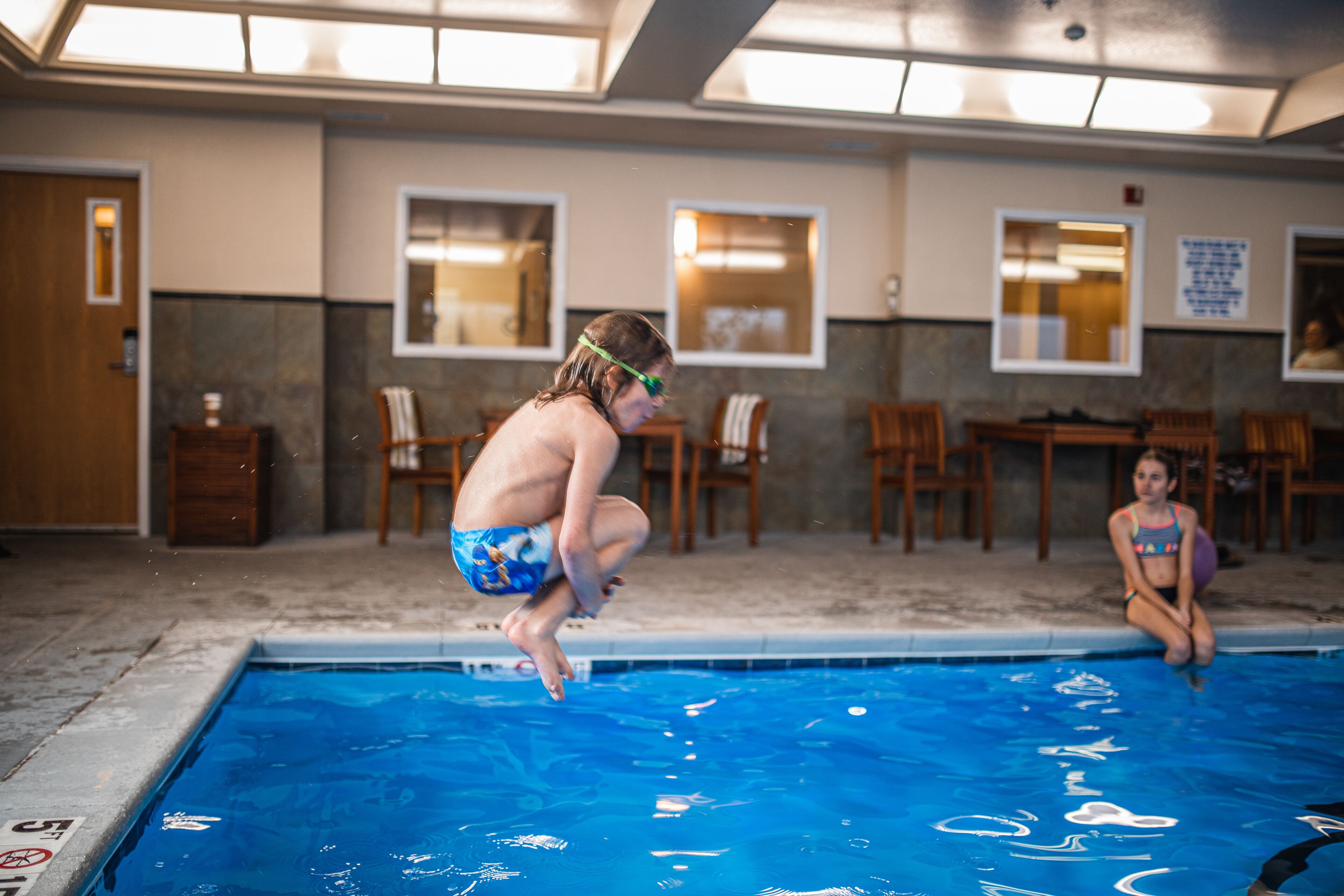 Una piscina con un trampolín, sillas y una mesa al fondo.