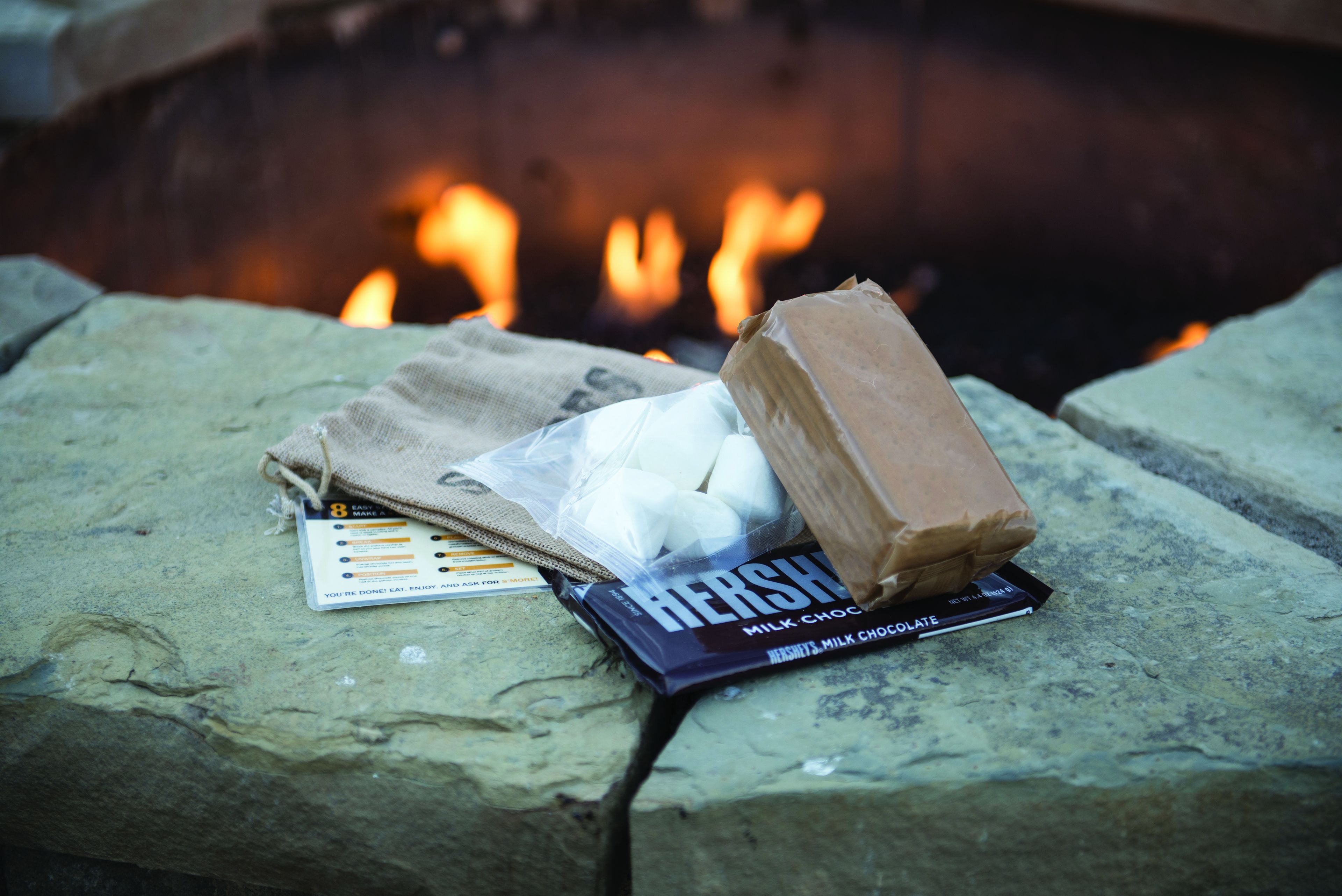 A Hershey's milk chocolate bar, marshmallows, and a card on a stone surface near a fire.