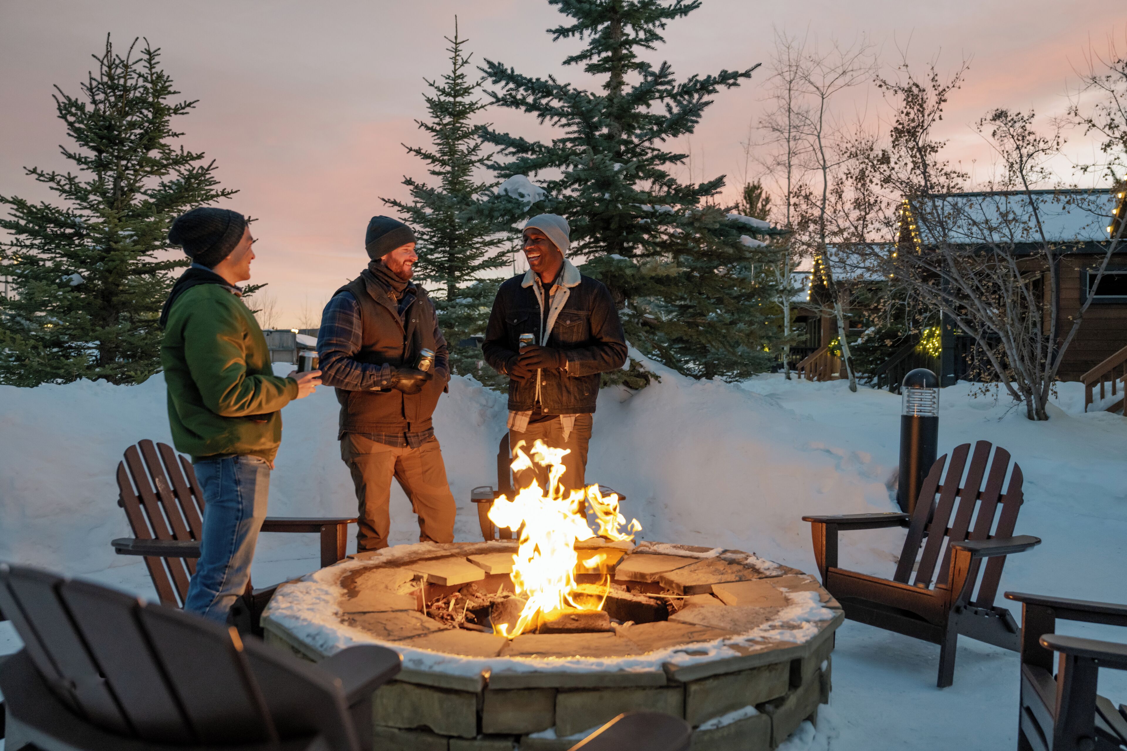 A group of people around a stone fire pit with chairs, set in a snowy outdoor area with trees and a building in the background.