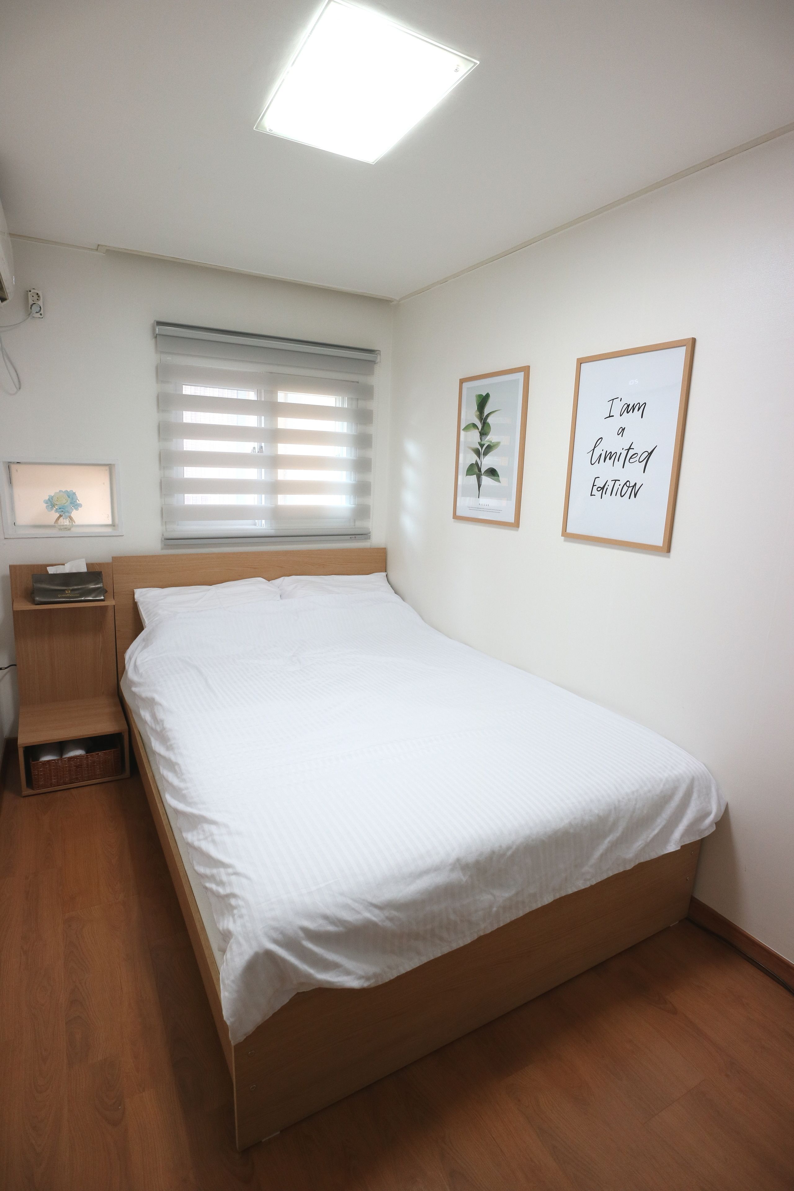 A neatly made bed in a room with a wooden floor, a bedside table, a nightstand, a window with blinds, and two framed pictures on the wall.