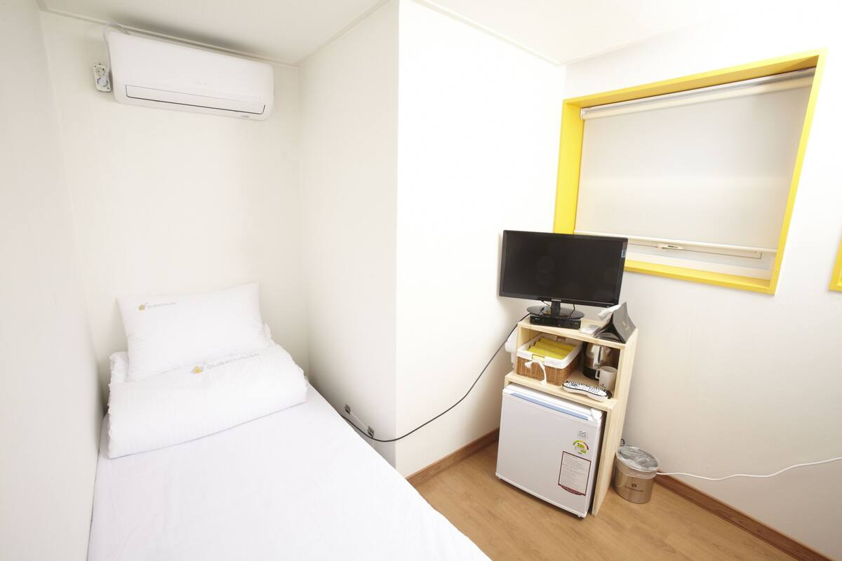 Habitació d'hotel amb un llit, una petita taula de treball amb un ordinador, una televisió i una finestra amb un marc groc.