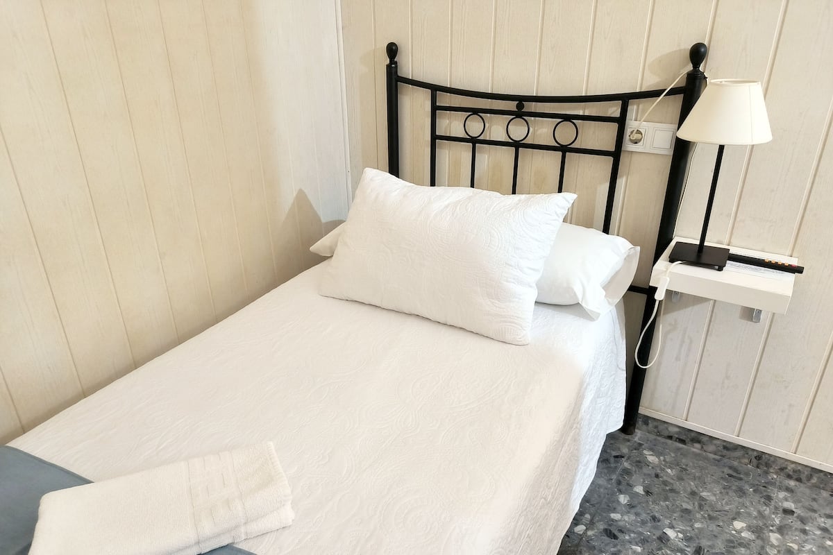 A neatly made bed with white linens and pillows, a bedside table with a lamp, and a wall-mounted power outlet.