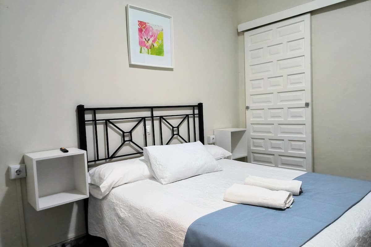 A neatly made bed with white bedding and a blue accent, a white nightstand with a lamp, and a white door with a geometric pattern.