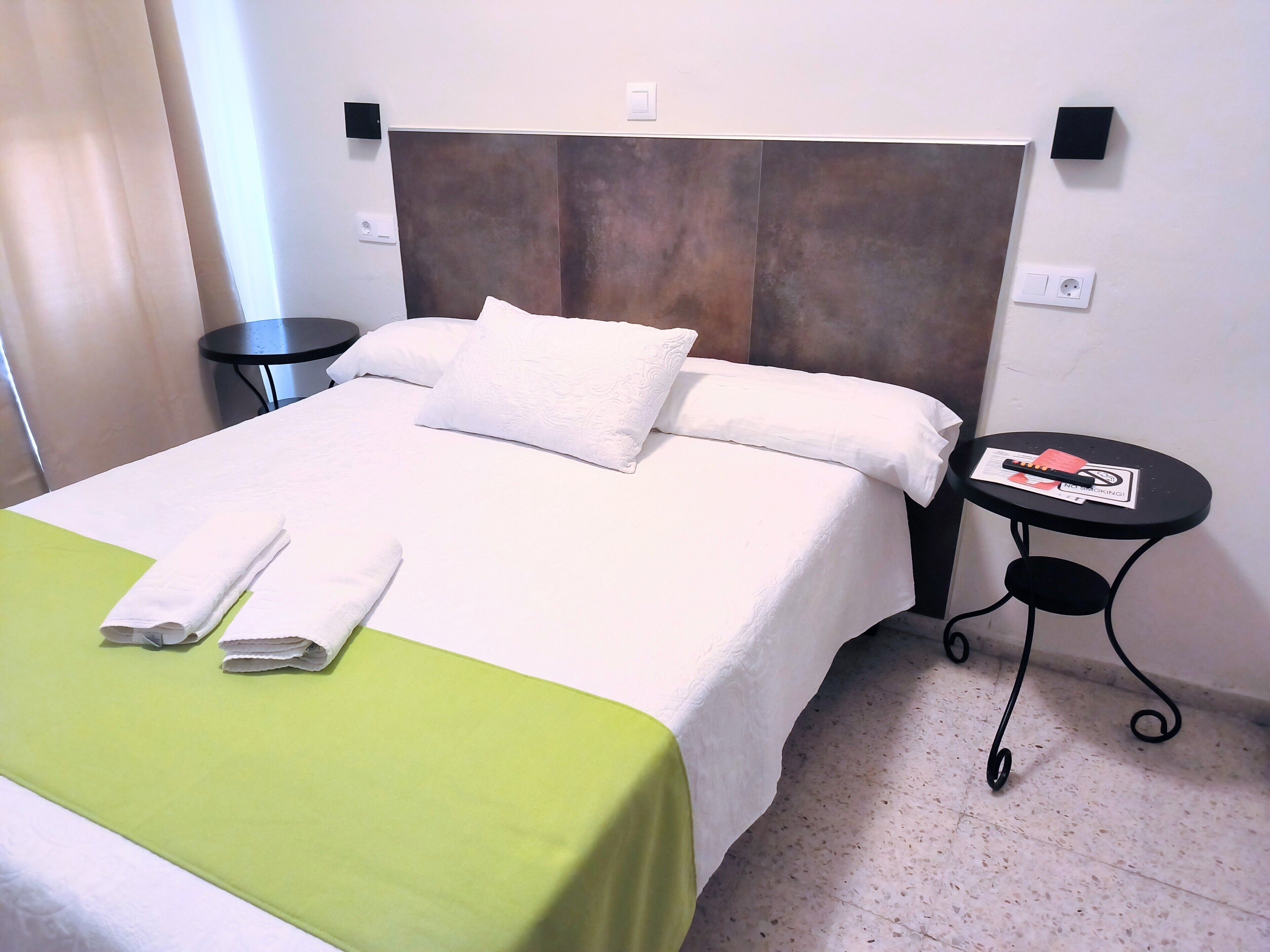 Una cama bien hecha con sábanas blancas y una colcha verde, una mesita redonda con un control remoto y un enchufe en la pared.