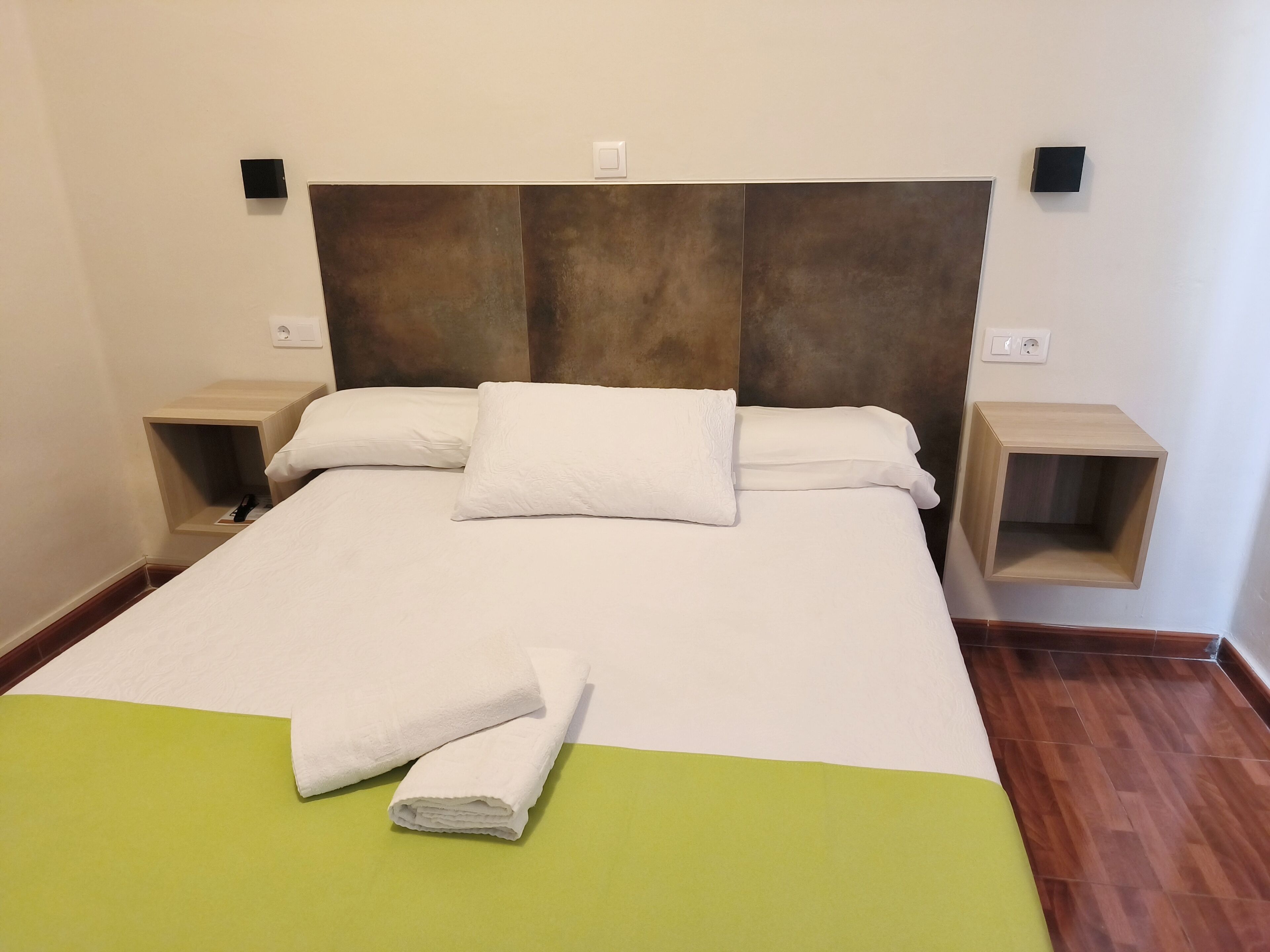 Un lit bien fait, avec du linge de lit blanc et une serviette pliée, une tête de lit en bois, des tables de chevet avec rangements et un élément décoratif vert sur le lit.