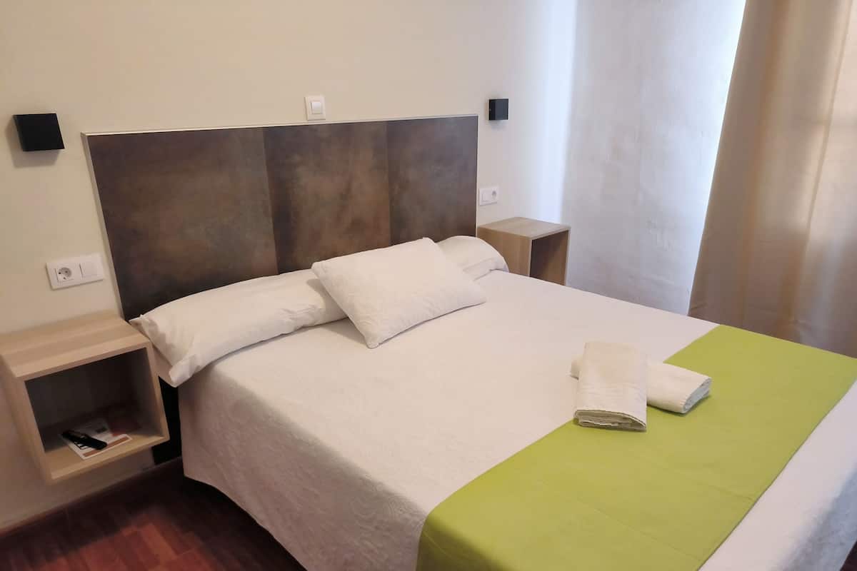A neatly made bed with white linens, a green throw blanket, and a folded towel on the footboard. There is a wooden nightstand with a remote control and a book beside the bed.