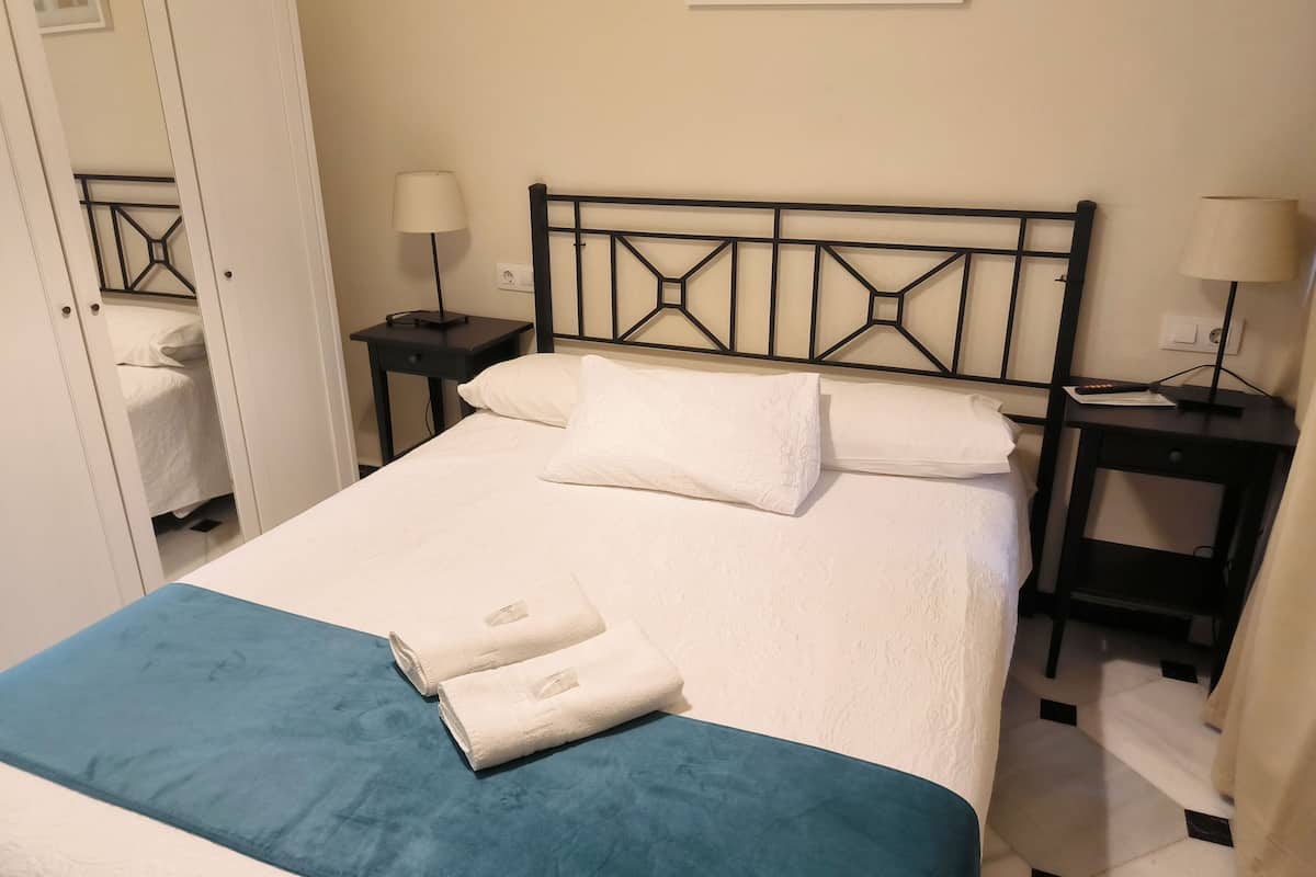 A neatly made bed with white linens and a teal blanket, flanked by two bedside tables with lamps, in a room with a simple, clean design.