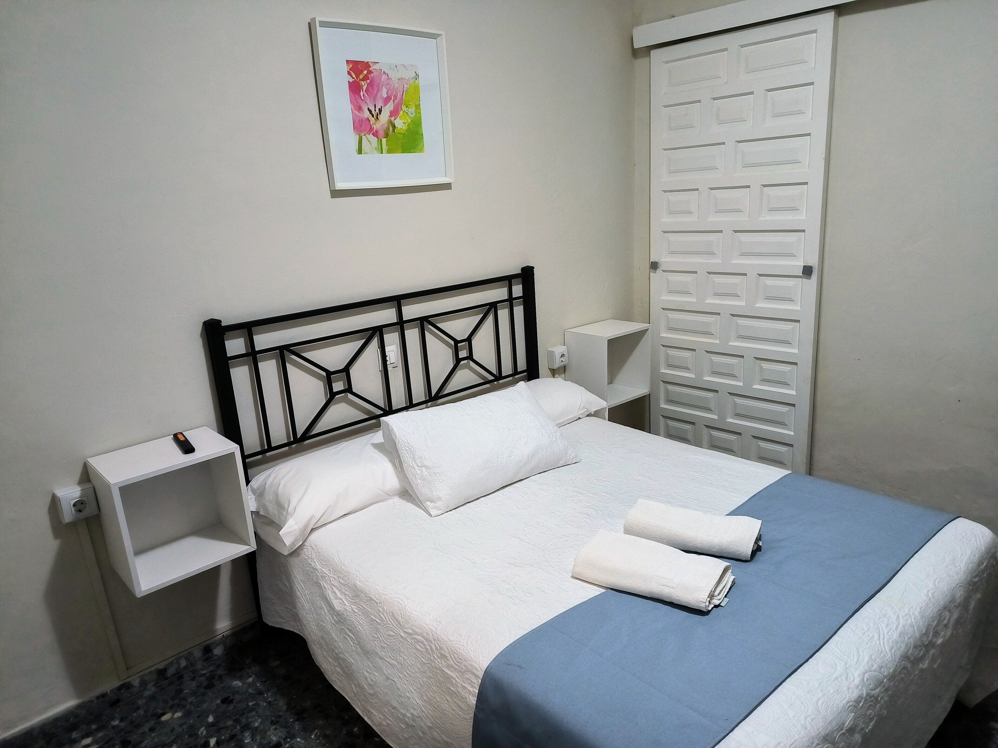 A neatly made bed with white linens, a blue blanket, and two rolled towels. There is a white shelf on the left and a white wardrobe with a geometric pattern on the right. A framed picture hangs on the wall above the bed.
