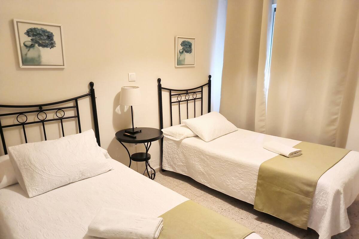 Two single beds with white linens, a small round table with a lamp, and two framed floral paintings on the wall.